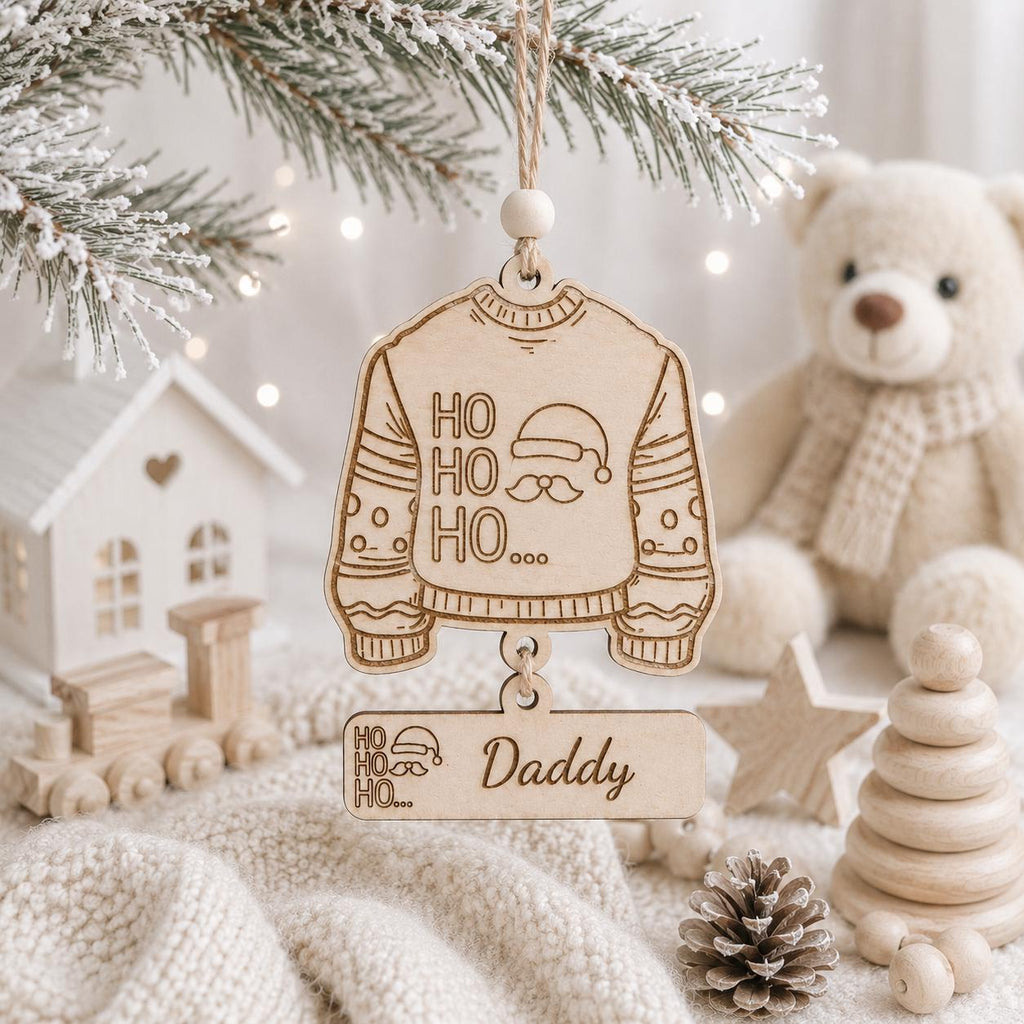 Personalized wooden Christmas ornaments hanging on a decorated tree branch, featuring a sweater design with 'HO HO HO' and a Santa hat, and a rectangular ornament engraved with 'Daddy' and a Santa hat. The scene is styled with a teddy bear, knitted blanket, and wooden toys.