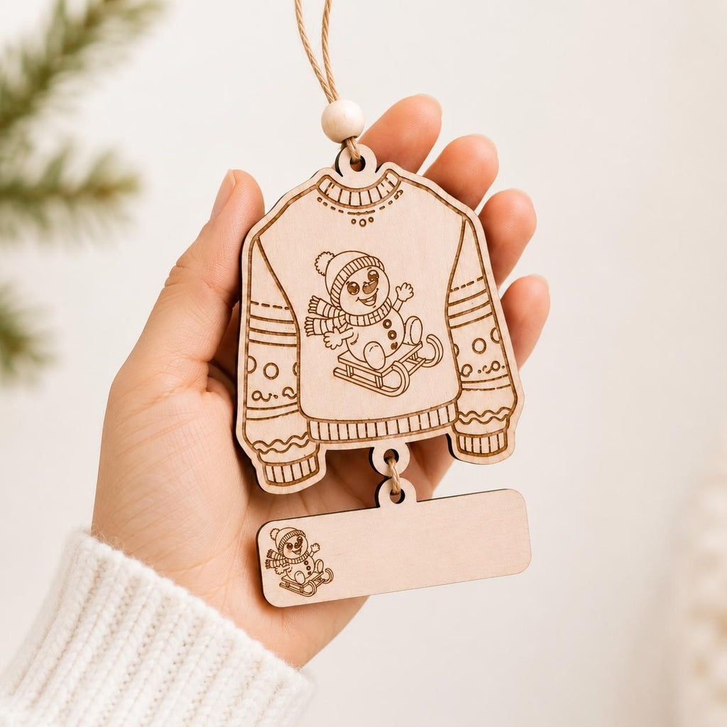 A hand holds a laser-cut wooden ornament shaped like a cozy sweater with a bear sitting on a sled, featuring a blank rectangular tag below it.